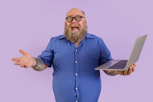 Joyful fat businessman in tight shirt with spectacles holds laptop on purple background