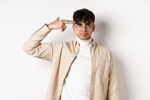 Annoyed young man making finger gun gesture on head, shooting himself from boredom or annoyance, tired of something stupid, standing on white background