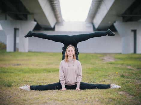 Two girls acrobats perform stand in the splits on the grass against the background of the bridge