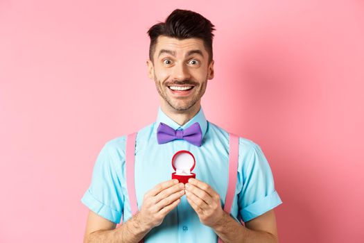 Valentines day. Romantic man in bow-tie showing engagement ring and smiling, asking to marry him, making proposal to lover on pink background