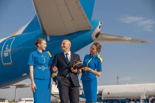 Excited pilot with two attractive stewardesses standing together in front of an airplane and smiling after landing