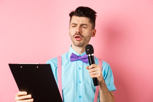 Holidays concept. Serious-looking man reading script from clipboard, holding microphone, entertain people on festive event, standing on pink background