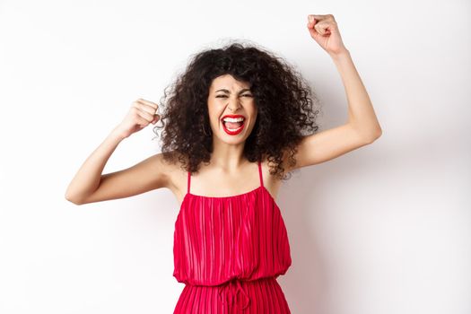 Cheerful emotive woman with curly hair, red dress, raising hands up and chanting, rooting for team, shouting wanting to win, standing on white background