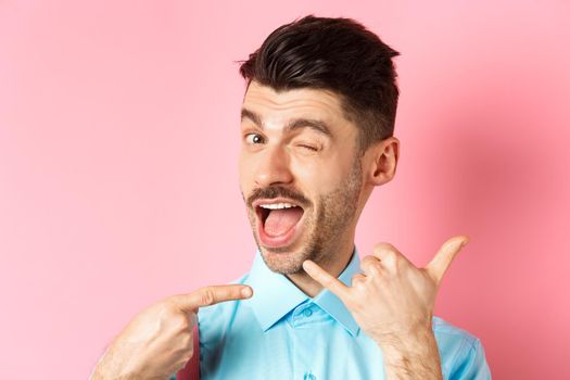 Cheeky and funny guy showing phone gesture, asking to call him and winking, telling dial his number, standing over pink background