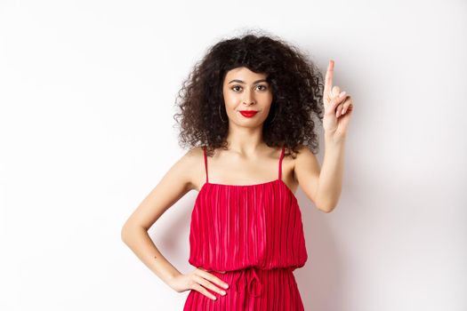 Smiling caucasian woman with curly hair, wearing red dress, showing rule number one gesture, raising finger and looking confident at camera, white background