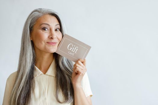Joyful grey haired Asian woman holds gift card posing on light grey background