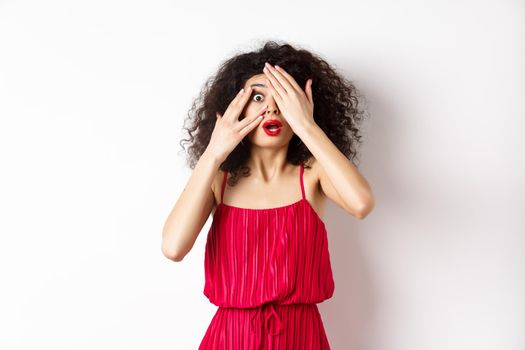 Excited curly-haired girl in dress, cover eyes with hands and peek through fingers, gasping shocked at camera, standing on white background