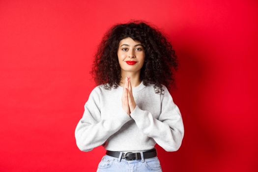 Namaste. Thankful young woman with curly hair and red lips show gratitude, smiling pleased at camera, standing on studio background.