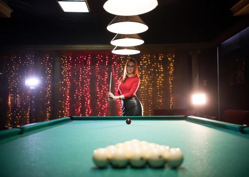 Billiard club. A woman with red hair standing by the table holding a cue