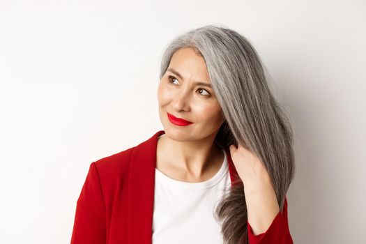 Beauty and haircare concept. Close up of elegant asian senior woman showing shiny and healthy grey hair, smiling and looking aside, white background
