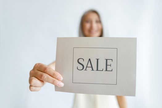 Positive mature woman stands in studio, focus on hand holding card with word Sale