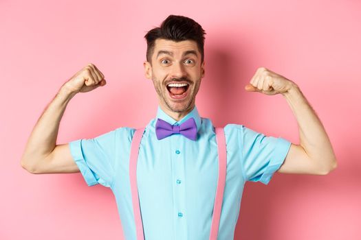 Smiling caucasian man with bow-tie and suspenders, showing muscles and feeling strong, flexing biceps to show-off, standing over pink background