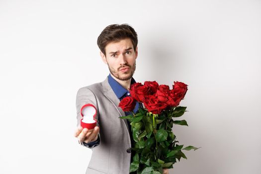 Handsome boyfriend in suit asking to marry him, standing with red bouquet of roses and engagement ring, looking pleading at camera, standing over white background