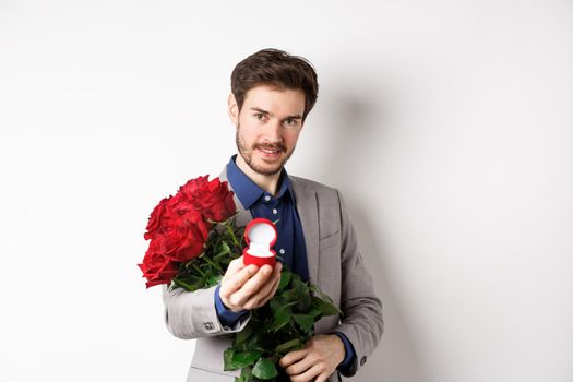 Romantic man with boquet of red roses asking to marry him, holding engagement ring and looking confident at camera, standing in suit over white background