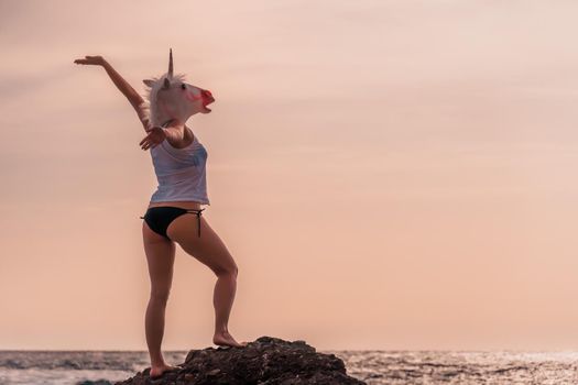 Unusual Woman in white t-shirt and black swimsuit with unicorn head mask dancing by the sea on the beach, warm sunset time strange funny weird photo.