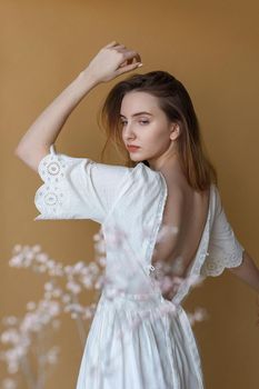 Beautiful young girl with long hair in white dress posing on beige background. White flowers on foreground out of focus