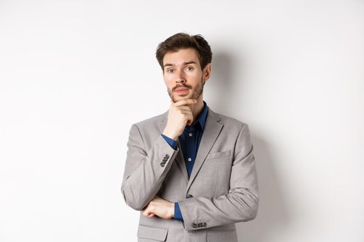 Thoughtful businessman in suit touching beard, thinking and looking at camera, deciding with pensive face, standing on white background