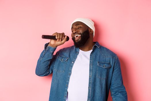 Handsome Black man in beanie and denim shirt singing karaoke, holding microphone, standing over pink background