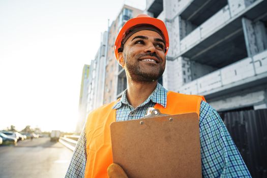 Young man engineer in workwear standing in construction site with clipboard