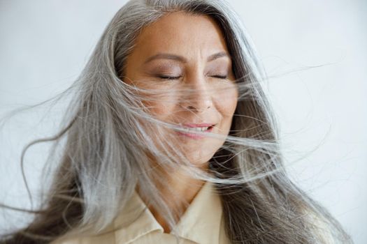 Happy mature Asian model with flying hoary hair stands on light grey background