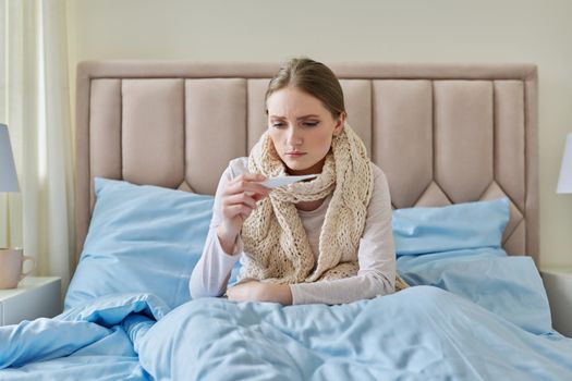 Sick young woman with thermometer in her hands, sitting at home in bed