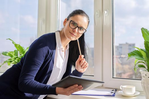 Business woman working near window using digital tablet, looking at camera