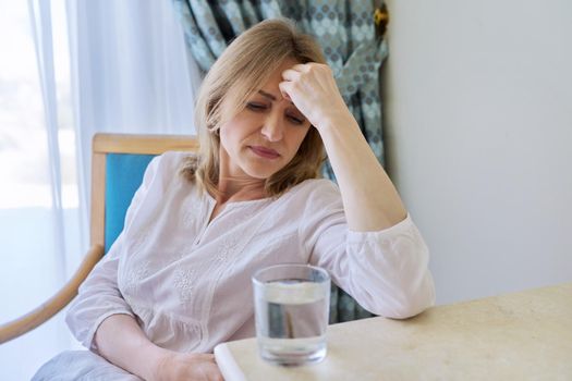 Headache, migraine, depression of mature woman sitting at home on chair with glass of water. Stress, health problems, illness, age people concept