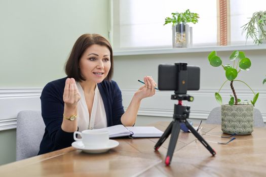Woman teacher, business, psychologist, blogger, lawyer talking online using smartphone. Female looking at phone webcam on tripod, background table with cup of coffee notepad, window