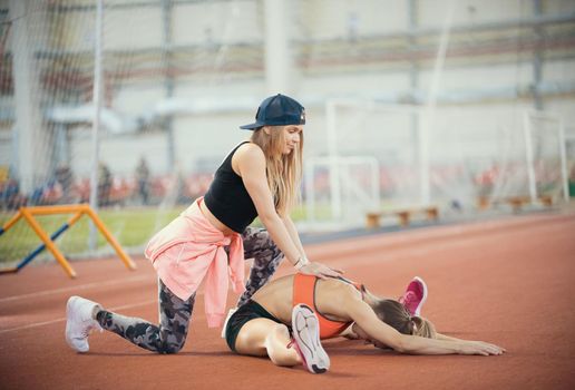 Young beautiful woman with blonde hair helps another woman to stretch her legs in the sports hall after training, side view