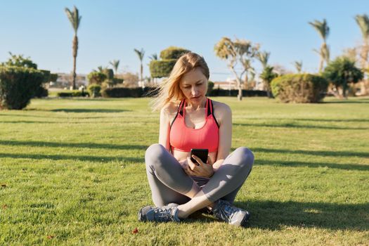 Mature woman doing morning exercises on green grass, with smartphone, video workout