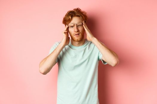 Portrait of redhead man in crooked glasses touching head and feeling dizzy or nauseous, having hangover or headache, standing over pink background