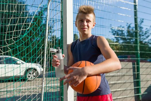 Teenager boy playing basketball with ball on the basketball court drinking water.