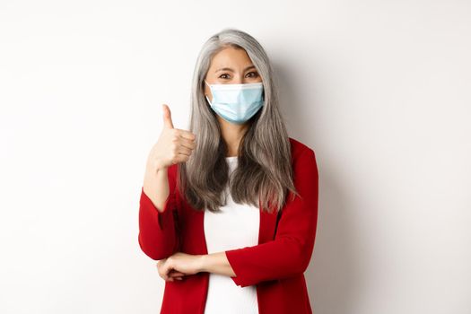 Coronavirus and business concept. Asian female entrepreneur in face mask looking cheerful, showing thumb-up in approval, praising using protective measures at work
