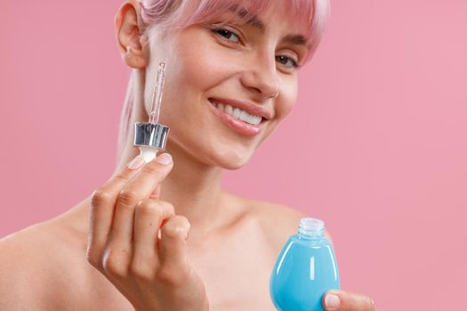 Portrait of young woman with pink hair holding dropper and a bottle of serum or hyaluronic acid, posing isolated over pink background