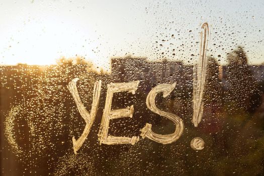 The word yes on the window with water drops on sunny background, yes written with white toothpaste