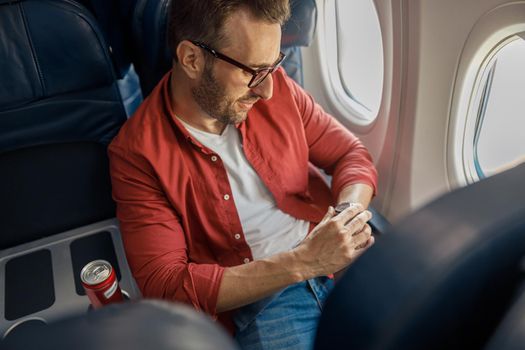 Handsome caucasian man in casual wear and glasses checking arrival time on his watch, sitting on the plane near the window