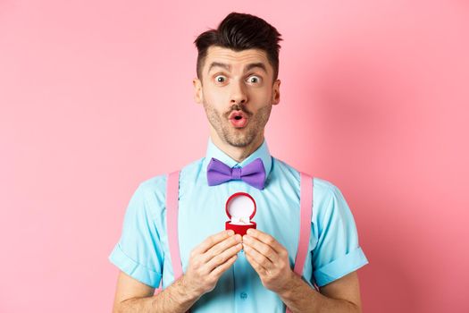 Valentines day. Funny man with moustache and bow-tie, looking excited and showing engagement ring, asking girlfriend to marry him, standing over pink background