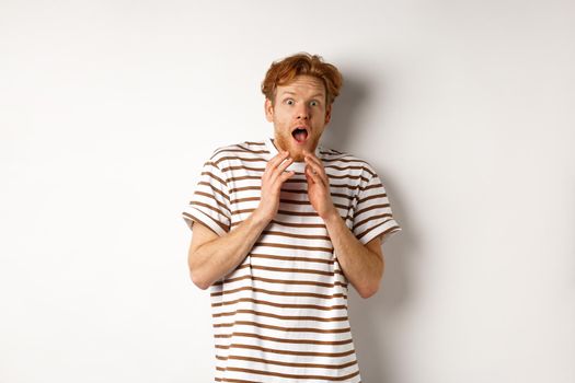 Timid young man with red hair, looking scared, jumping and screaming from fear, standing over white background