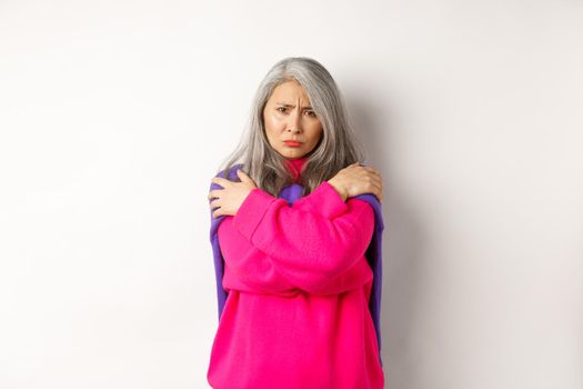Defensive and offended asian middle-aged woman hugging body, comforting herself and looking angry at camera, frowning insulted, standing over white background