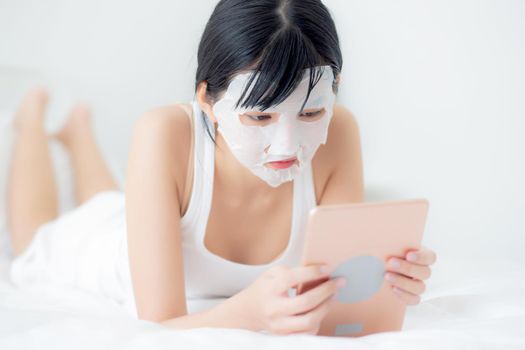 Beautiful young asian woman with sheet facial mask lying watching tablet computer on bed at bedroom, beauty girl applying face mask for skincare of wrinkle at home, skin care, health and wellness.