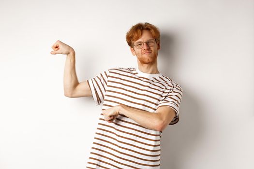 Image of confident and strong redhead man flexing biceps, showing muscles after gym, standing over white background