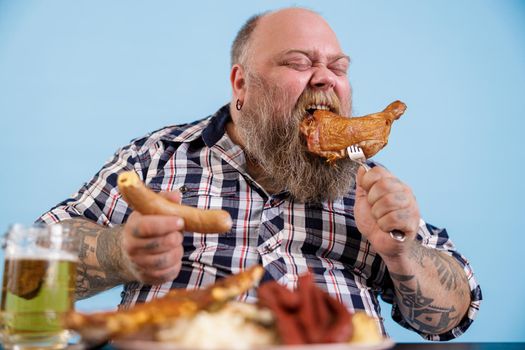 Expressive person with overweight eats chicken leg at table with greasy food