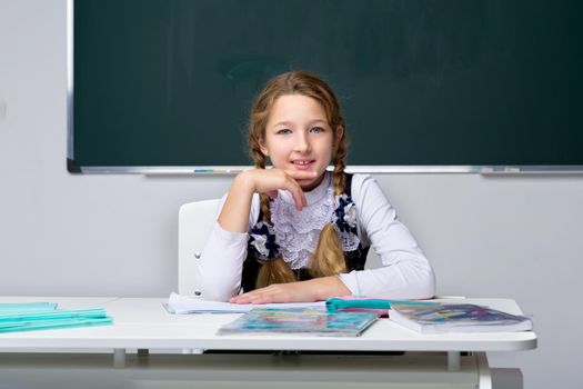 Portrait of a cute schoolgirl. A pensive, tired girl sits at a desk. Elementary school student posing in front of the blackboard. Back to school, education concept.