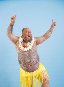 Jouful plump man with bare chest in yellow grass skirt a has fun on light blue background