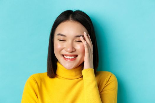 Beauty and makeup concept. Close up of beautiful asian woman laughing and looking flushed from compliments, standing over blue background