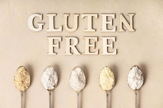Metal spoons of various gluten free flour (almond flour, oatmeal flour, buckwheat flour, rice flour, corn flour) and gluten free lettering made of wooden letters, flat lay, top view