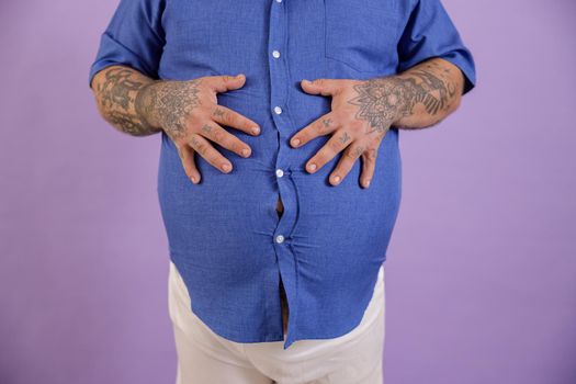 Man with overweight and tattoos holds hands on large belly on purple background