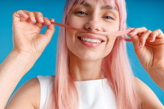 Close up portrait of happy young woman with natural long pink dyed hair holding a strand of hair as a moustache and smiling at camera, posing isolated over blue studio background