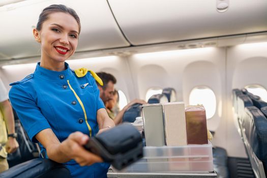 Portrait of charming airline stewardess smiling at camera, holding terminal for passenger to make a payment