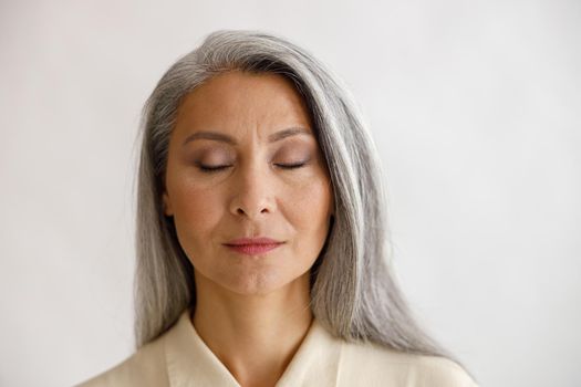 Tranquil grey haired lady with closed eyes and elegant makeup poses on light background